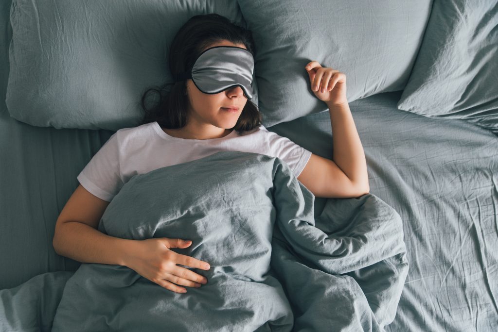 Woman Sleeping On Gray Sheets With Gray Sleep Eye Mask On.