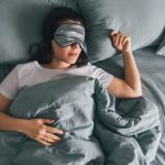Woman Sleeping On Gray Sheets With Gray Sleep Eye Mask On.