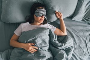 Woman sleeping on gray sheets with gray sleep eye mask on.