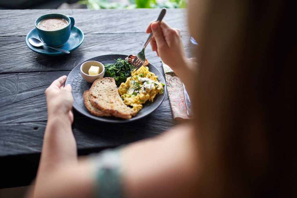 Closeup Shot Of Delicious Brunch Plate With Multi-Grain Bread, Butter, Fried Greens, Egg, Organic Coffee Being Eaten With Fork On Rustic Table Near Window