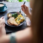 Closeup Shot Of Delicious Brunch Plate With Multi-Grain Bread, Butter, Fried Greens, Egg, Organic Coffee Being Eaten With Fork On Rustic Table Near Window
