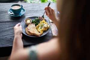 Closeup shot of delicious brunch plate with multi-grain bread, butter, fried greens, egg, organic coffee being eaten with fork on rustic table near window