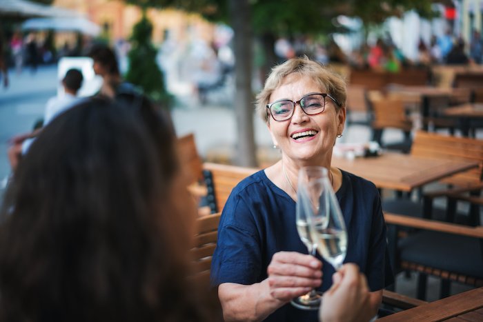 Beautiful happy older woman sitting with her friend in a street cafe clinking glasses with champagne