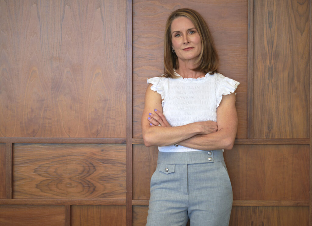 Adrien Cotton In Gray Slacks And A White Blouse, Leaning Against A Wood Paneled Wall With Arms Crossed.