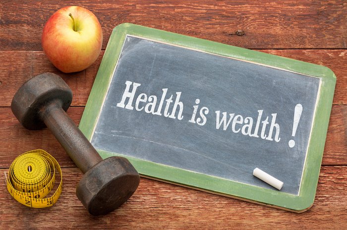 Health Is Wealth Written On A Slate Blackboard Sign Against Weathered Red Painted Barn Wood With A Dumbbell, Apple And Tape Measure