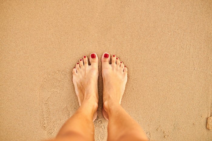 Bare Feet Standing On A Sandy Beach With Red Toenails.