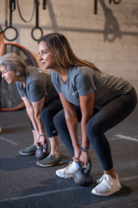 Two Middle Aged Women In A Strength Gym Doing Deadlifts With Kettlebells.