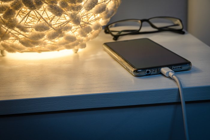 Smart Phone Charging With Usb Cable On A White Nightstand With A Soft Light Next To It