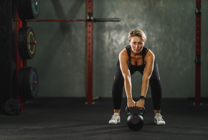 Woman Faces The Camera While Preparing To Lift A Kettlebell Off Of The Floor.