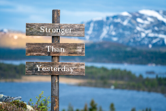 Stronger Than Yesterday Text On Signpost Outdoors During Blue Hour And Sunset. Snow Capped Mountains, Lake And Forest In The Background.