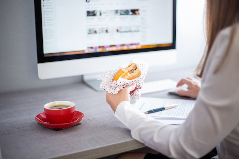 Young Business Woman Eating A Fast Food Burger With Coffee At The Workplace In Front Of A Computer Screen.