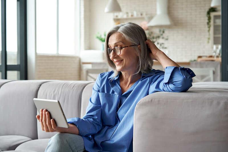 Middle Aged Adult Woman Sitting On The Couch And Holding A Digital Tablet.