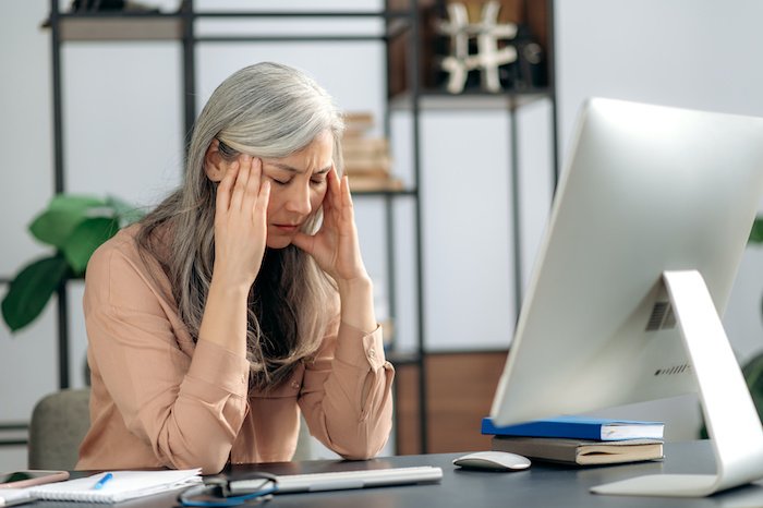 Tense Older Woman Sitting At A Desk With Her Hands On Her Temples And Eyes Closed