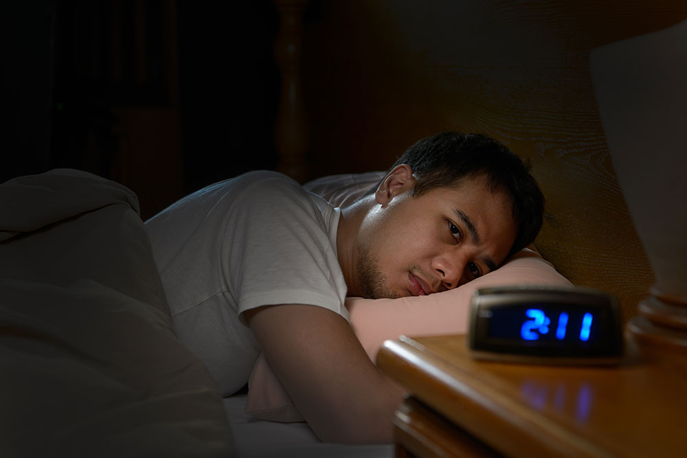 Person In A White T-Shirt Lying In Bed In The Dark Looking At A Clock On The Bedside Table.