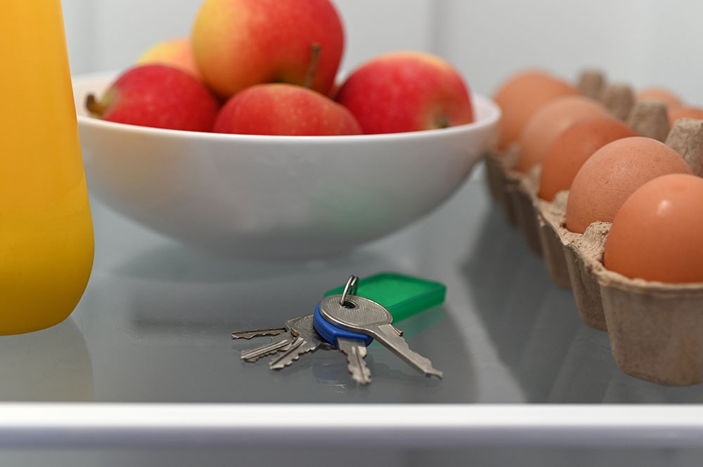 Close Up Of A Shelf In The Refrigerator. There Is A Set Of Keys Sitting On The Shelf In Front Of A Bowl Of Fruit And Next To A Carton Of Eggs.